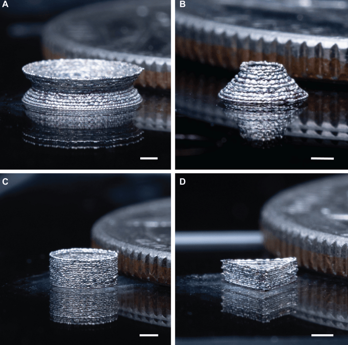 使用 Meta-NFS 逐层沉积法打印的 3D 结构照片。( A ) 银双锥结构。( B ) 银截锥体。( C ) 银环。( D ) 银三角棱柱。这些结构与一枚美国 25 美分硬币（直径 24.26 毫米，厚度 1.75 毫米）并排放置。比例尺：1 毫米。
