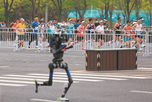 4月19日，参赛的长跑选手与杭U赛队自主人形机器人选手宇树H1（前）在比赛中。