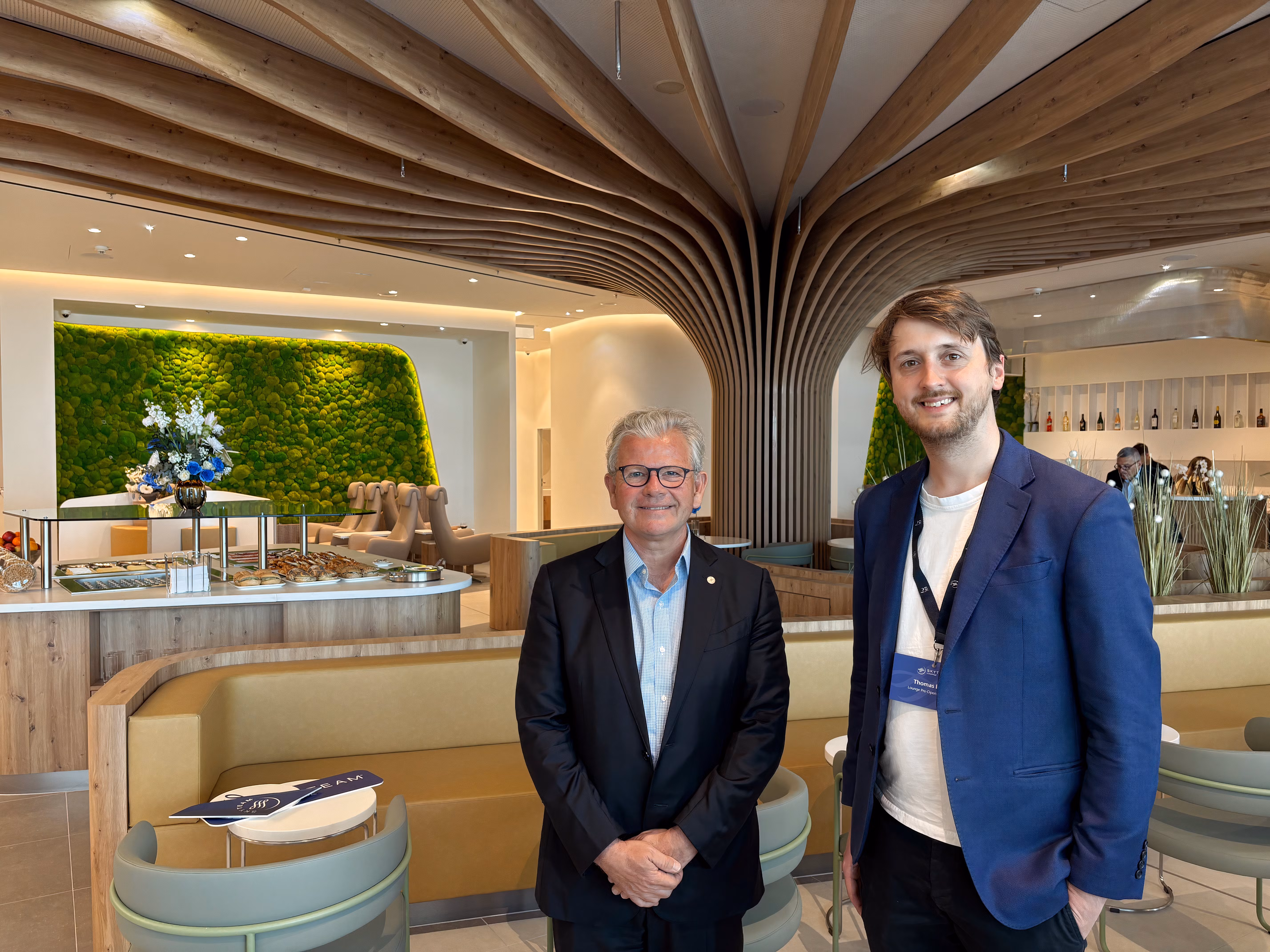 两名男士，帕特里克·鲁（左）和汤姆·布恩（右），在现代的天合联盟贵宾室中站在一起。贵宾室设有一个引人注目的建筑中心，弯曲的木条像树冠一样在天花板上展开。背景中有一个自助餐区，提供各种糕点，以及一面充满活力的绿色苔藓墙，为现代设计增添了自然元素。