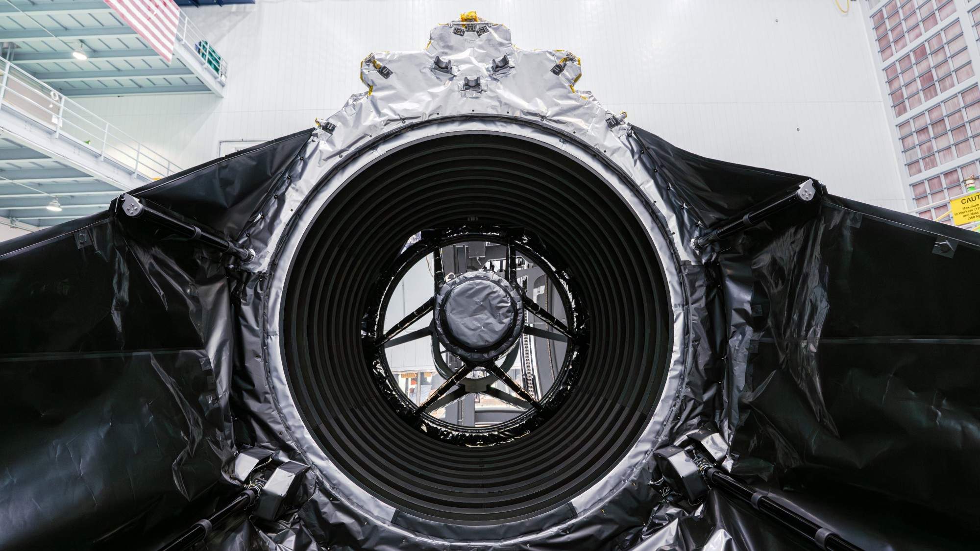 Technicians in clean suits stand in a hangar next to a large rectangular spacecraft wrapped in metal foil