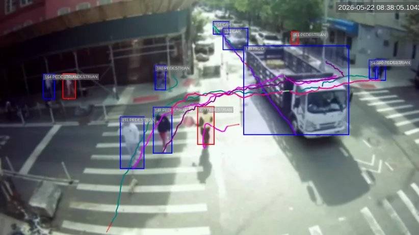 Vista aérea de um cruzamento movimentado de cidade, mostrando sobreposição de detecção por IA: caixas delimitadoras azuis e vermelhas identificam pedestres, carros e um caminhão rígido, com linhas de trajetória coloridas rastreando seus movimentos na cena. O carimbo de data/hora no canto superior direito mostra 2026-05-22 08:38:05.