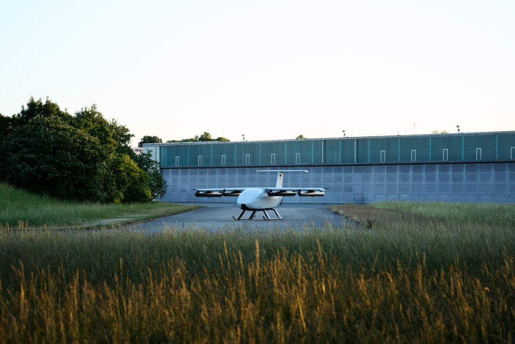 Petit drone blanc avec pales de rotor, stationné sur une piste devant un hangar bleu, avec des herbes hautes au premier plan.