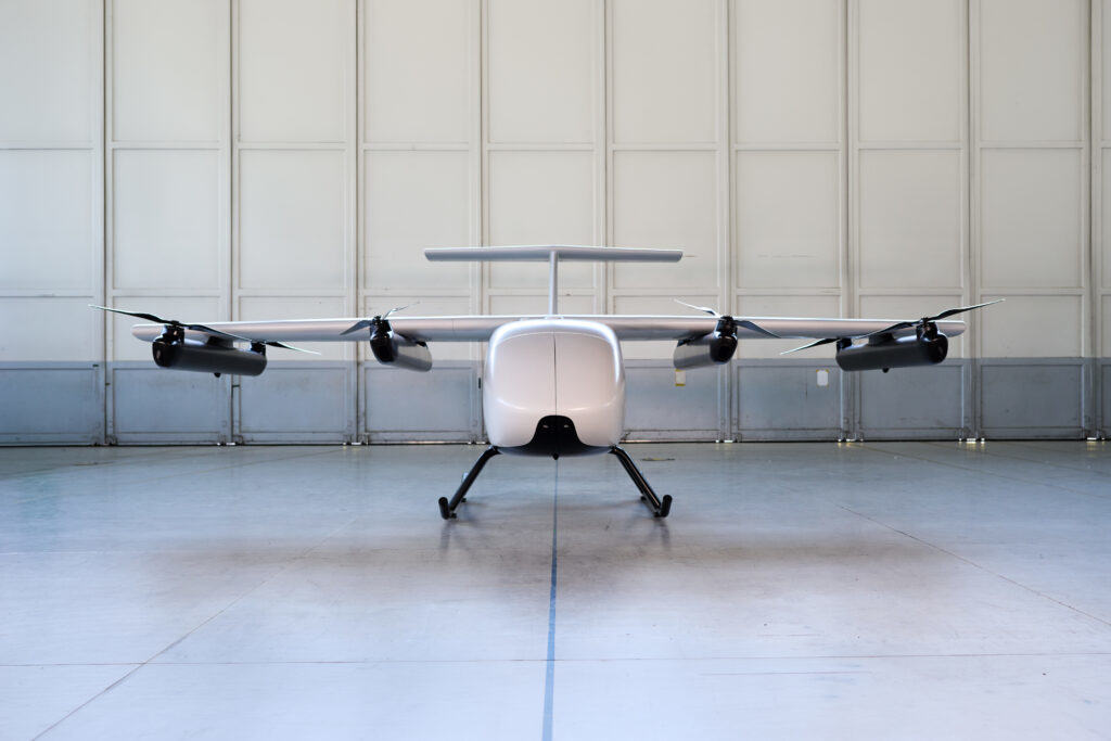Vue de face d’un petit avion bimoteur blanc stationné dans un hangar, avec une grande porte blanche en arrière-plan.