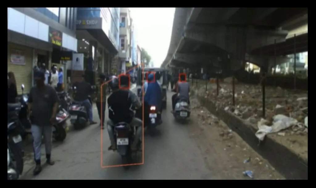 印度城市道路上摩托车手的街景摄像头画面，计算机视觉检测框用橙色高亮显示骑手和头盔区域。