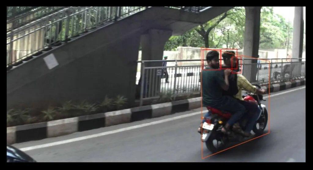 道路摄像头画面:两名骑手共乘一辆摩托车经过人行天桥下方,橙色和红色检测框高亮显示骑手及其未受保护的头部。