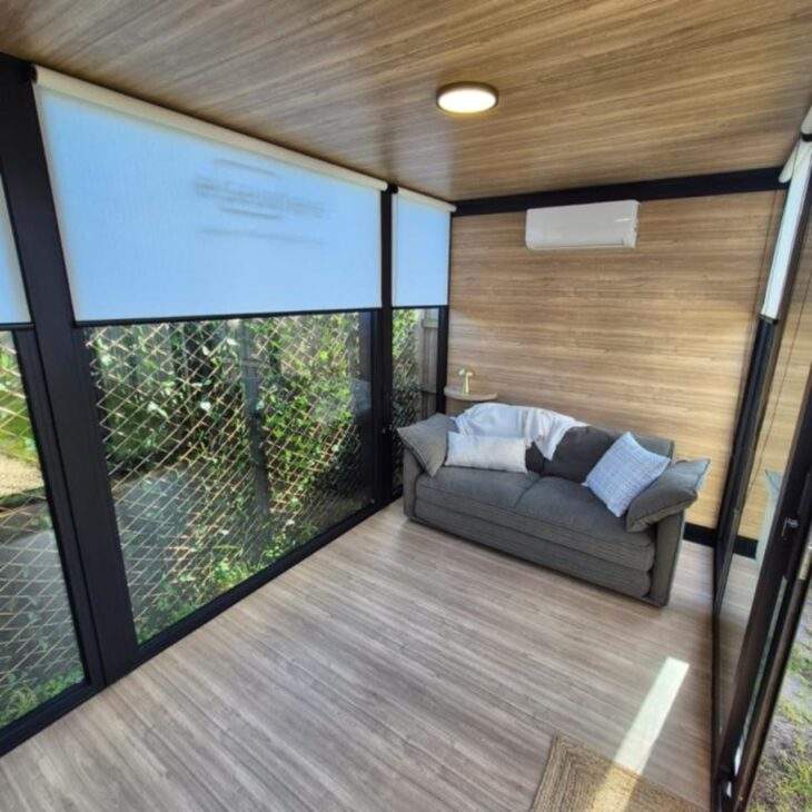 Interior de un módulo desmontable a la venta en Australia, que parece una habitación ya amueblada con sofá, aire acondicionado y grandes ventanales de vidrio. El modelo más pequeño cuesta aproximadamente 94.000 reales brasileños, mide 2,7 m x 2,4 m y se puede ensamblar en el patio trasero en pocos días.