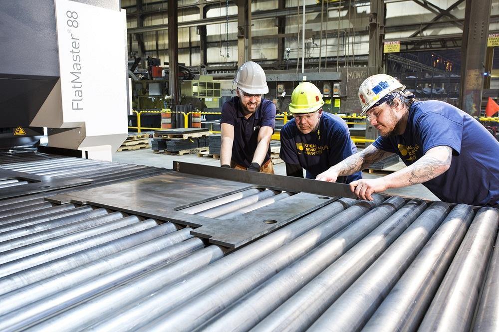 Trois personnes vérifient si les tôles métalliques sur le tapis roulant sont planes.