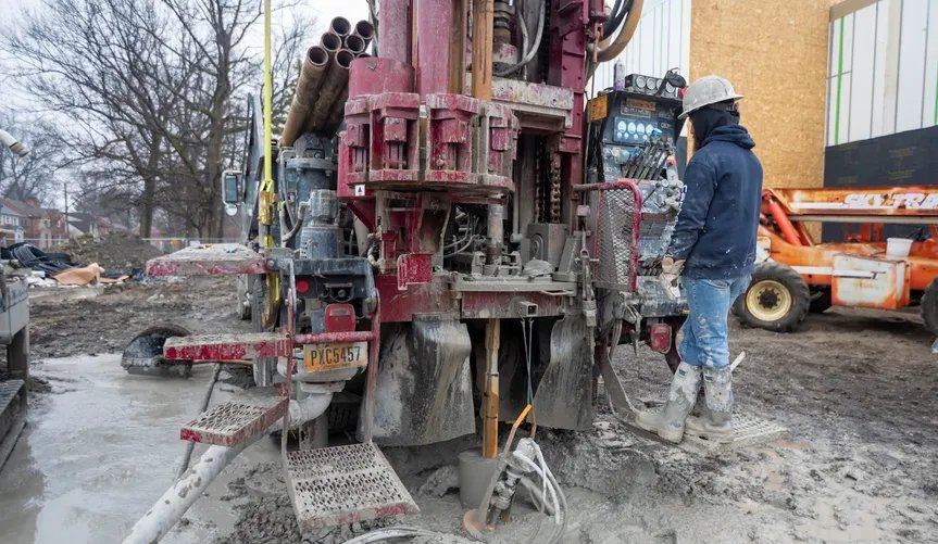 Workers operating a red drilling rig on muddy ground, wearing jeans, blue jackets, and hard hats