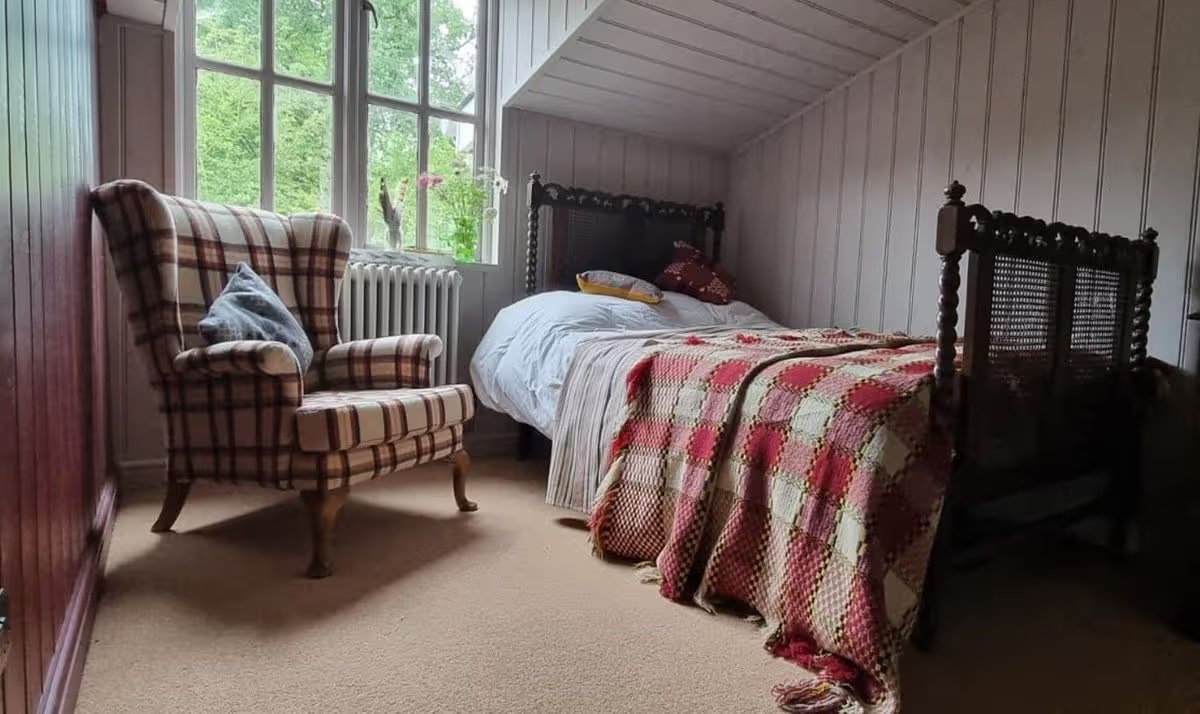 Jane Beck Welsh Blankets in ihrem renovierten Zuhause in der Nähe von Tregaron, Ceredigion. Dies ist eines von fünf Schlafzimmern mit einem traditionellen dunklen Holzbett, rostroten und cremefarbenen traditionellen walisischen Decken, hellgrauer Holzvertäfelung und einem Sessel am Fenster. Die Renovierung dauerte 29 Jahre, und sie hatte anfangs nur 23 Pfund in der Tasche.