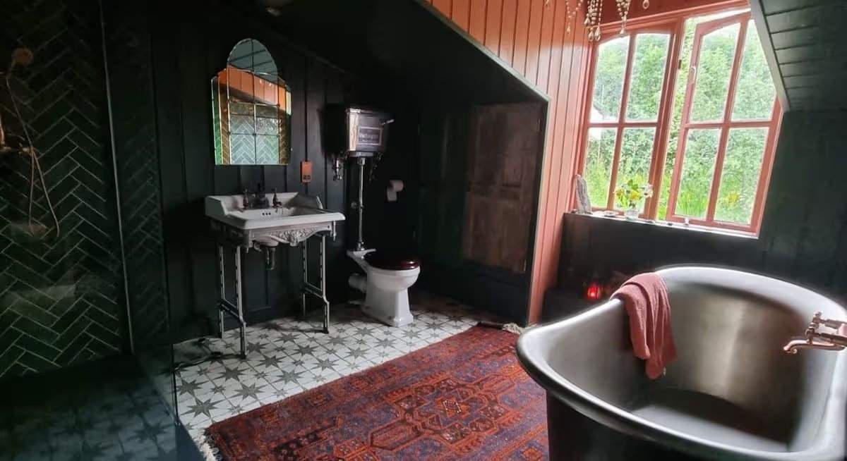 Das Badezimmer verfügt über eine freistehende Zinkbadewanne, eine begehbare Dusche mit dunkelgrünen Fliesen, eine traditionelle Toilette und ein Waschbecken sowie eine Holzvertäfelung in Schwarz, Grau und zartem Pfirsich. Jane Beck Welsh Blankets in ihrem renovierten Zuhause in der Nähe von Tregaron, Ceredigion. Die Renovierung dauerte 29 Jahre, und sie hatte anfangs nur 23 Pfund in der Tasche.