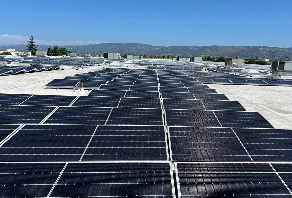 A large rooftop with numerous solar panels; hills and blue sky in the distance