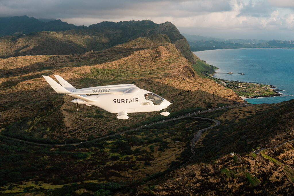 A small white Surf Air aircraft, with tail number N401NZ, flies over rugged coastal hills, with a winding road passing below and the blue ocean stretching along the right coast.