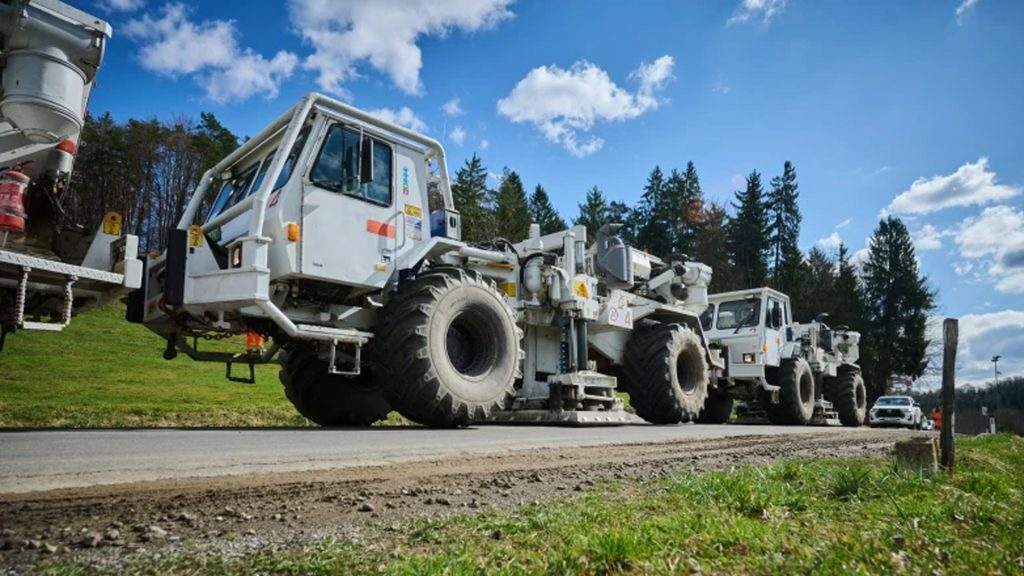 Ряд больших белых промышленных грузовиков с огромными шинами, припаркованных у дороги, на фоне голубого неба, редких облаков и деревьев.