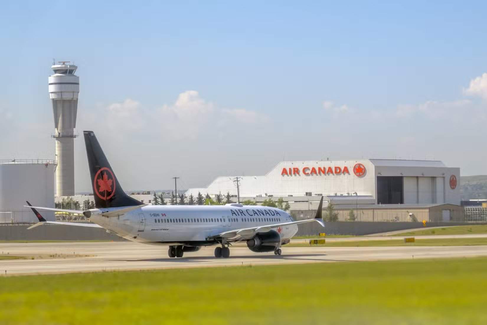 Un Boeing 737 MAX 8 d’Air Canada roule sur la piste, avec en arrière-plan la tour de contrôle et le hangar d’Air Canada