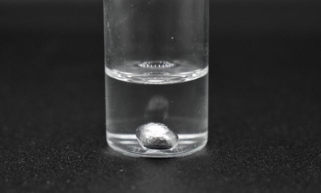 Foto eines kleinen Glasbehälters mit einer klaren Flüssigkeit und einem Tropfen Galliummetall am Boden.