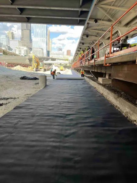 Workers unroll black geocomposite membrane on a gravel track bed next to an elevated railway platform, with excavation equipment and the Chicago skyline in the distance.