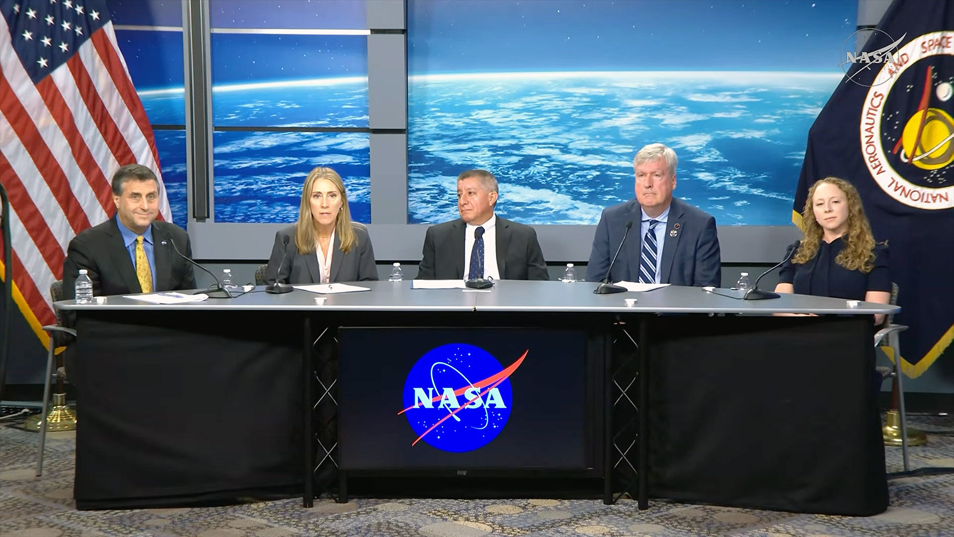 Две женщины и трое мужчин в деловой одежде сидят за столом с логотипом NASA на пресс-конференции