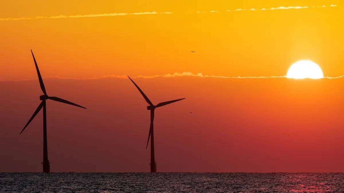 Au coucher du soleil, la surface de la mer reflète la lumière du soleil, les turbines d'un parc éolien offshore se découpent en silhouette dans un coucher de soleil dégradé de rouge.